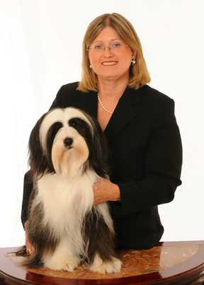 Blond woman in dark suit and pearl necklace standing behind black-and-white Tibetan Terrier sitting on inlaid wooden table
